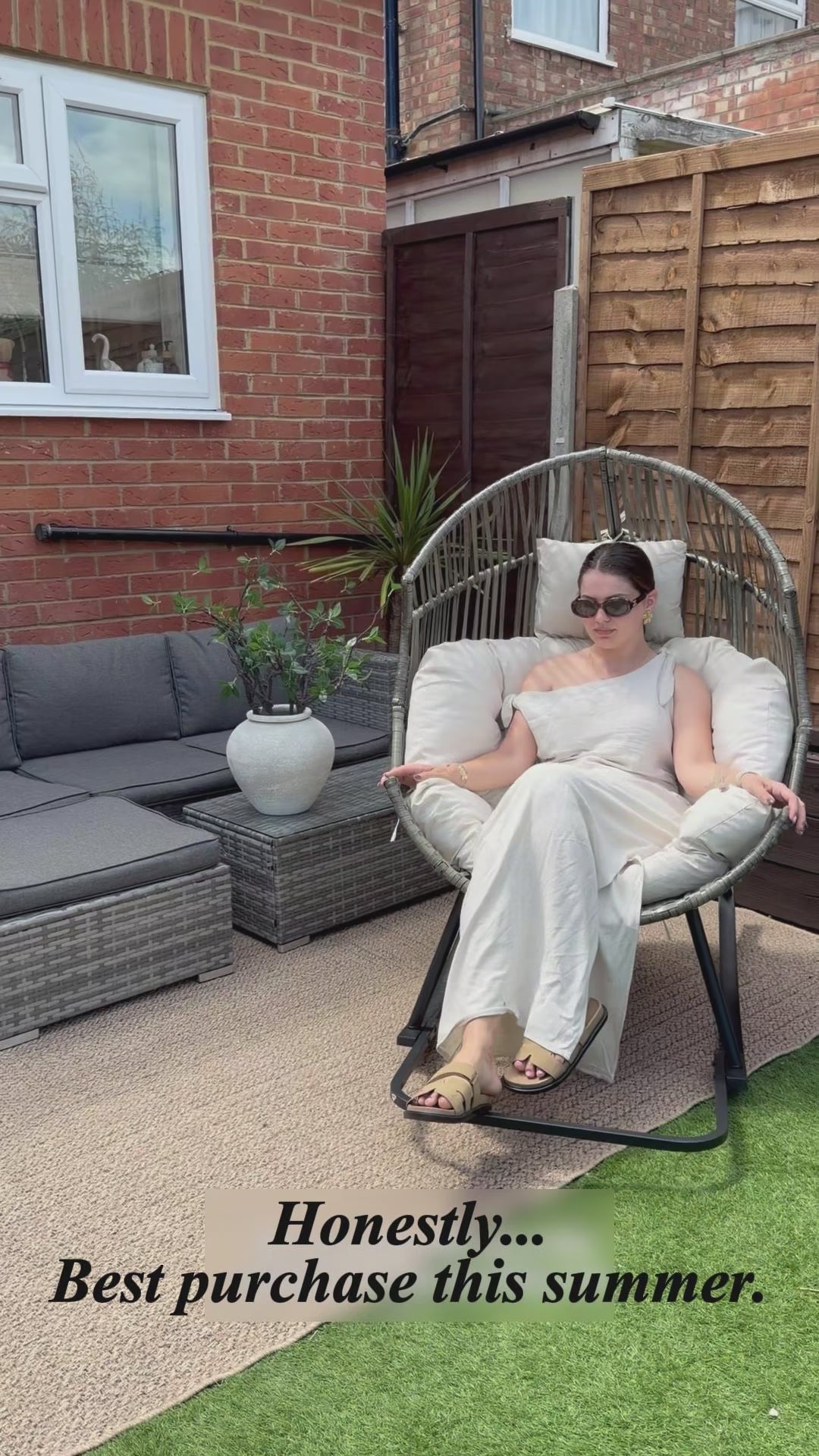 Woman demonstrating the Oversized Egg Rattan Rocking Chair in a modern British garden, showing its spacious seat and smooth rocking motion.
